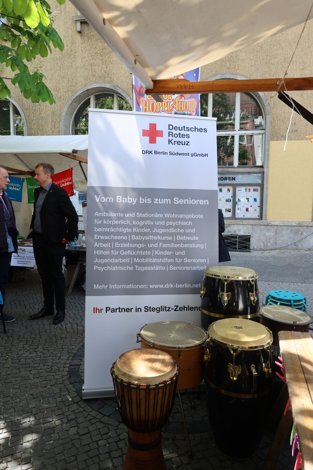 Infostand vom DRK Berlin SüdWest. Im Vordergrund sind die Trommeln gruppiert, im Hintergrund steht Herr Steinhoff, Bezirksstadtrat Stadtentwicklung.