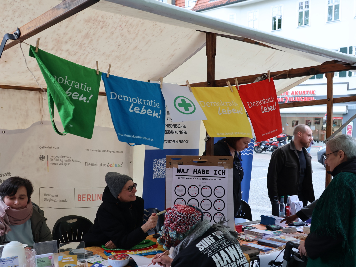 Infostand der Partnerschaft für Demokratie. Um den Stand herum stehen und sitzen mehrere verschiedene Menschen. Es gibt eine Stoffgirlande aus Quadraten in verschiedenen Farben: grün, blau, gelb, rot. Auf jedem steht in weißen Buchstaben "Demokratie leben". Dazwischen hängt ein Zeichen mit einem grünen Kreuz umrandet von einem grünen Kreis, das Zeichen für erste Hilfe Station. Im Hintergrund ist der Stand des Sozialpsychiatrischen Dienstes erkennbar.