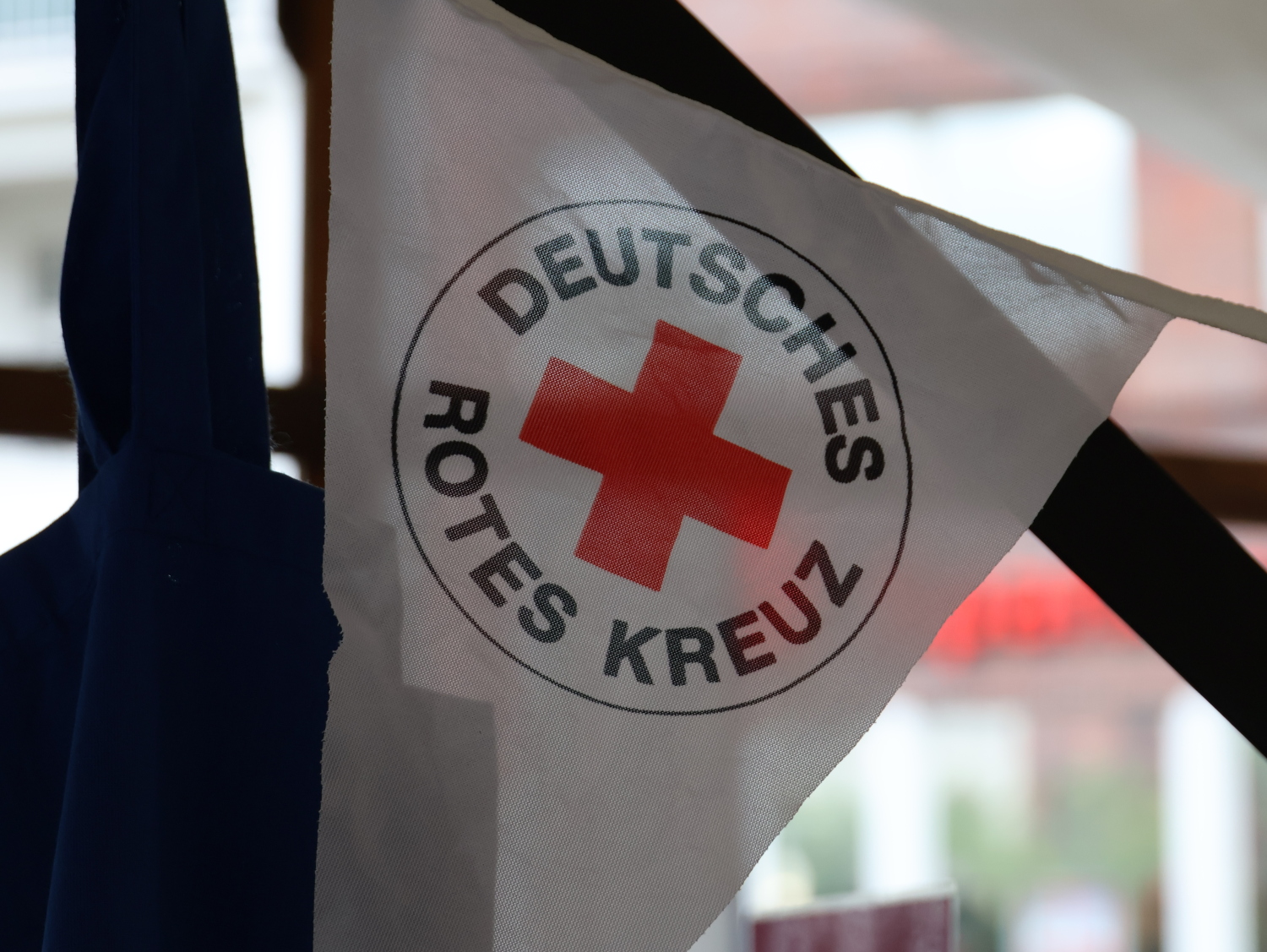 Infostand vom Deutschen Roten Kreuz. Es ist nur ein Wimpel der Girlande abgebildet. Der Wimpel ist weiß und trägt das Logo des DRK: ein großes rotes Kreuz, über dem Kreuz in schwarzer Großbuchstaben DEUTSCHES unter dem Kreuz in schwarzen Großbuchstaben ROTES KREUZ. Alles ist von einem dünnem schwarzem Kreis umrandet.