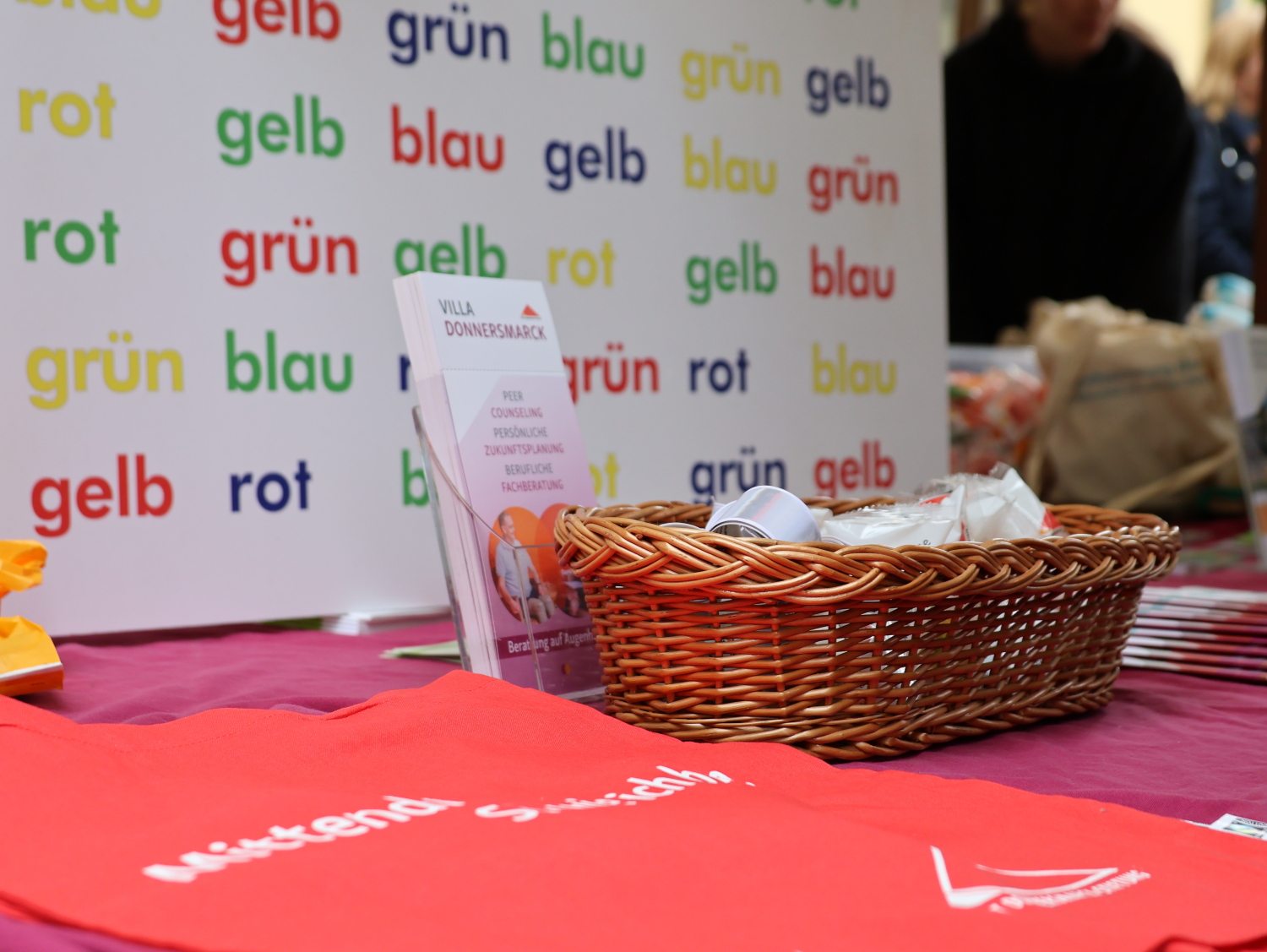 Infostand der Villa Donnersmarck. Es wurde nur ein Ausschnitt abgebildet. Im Hintergrund ist ein Schild zu erkennen auf dem Farbnamen aufgeschrieben sind: gelb, grün, blau, rot, aber immer in einer anderen Farbe. Es ist ein Farbtest. Im Vordergrund ist ein geflochtenes Körbchen zu erkennen, Flyer und rote Beutel der Villa Donnersmarck.