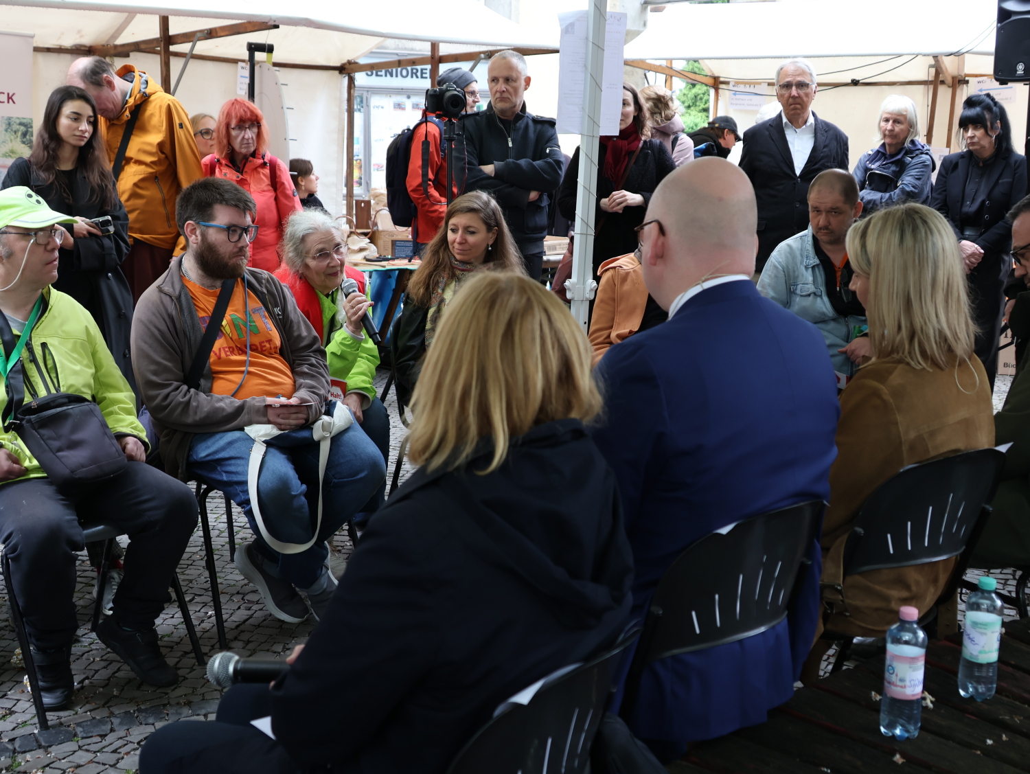 Politik-Talk "Mitreden! Inklusion im Bezirk" mit Blick auf die Fragestellenden und interessierte Zuhörer/innen. Einige tragen bunte und leuchtende Kleidung, andere eher dunkle Sachen.Die Stadträtinnen und Stadträte sind von hinten zu sehen.