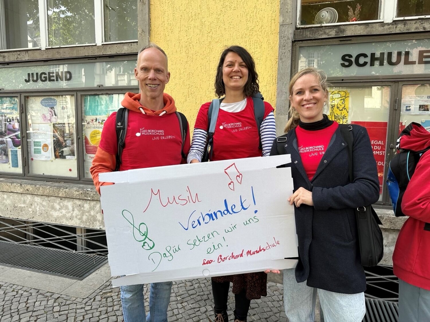 3 Mitarbeiter*innen der Leo-Borchard-Musikschule nehmen teil. Sie tragen rote Erkennungsshirts. Sie halten gemeinsam ein Demoschild auf dem steht in verschiedenen Farben (rot) Musik (blau) verbindet! (grün) Dafür setzen wir uns ein! (rot) Leo-Borchard-Musikschule. Es gibt dazu einen grünen Notenschlüssel und rote herzförmige Noten.