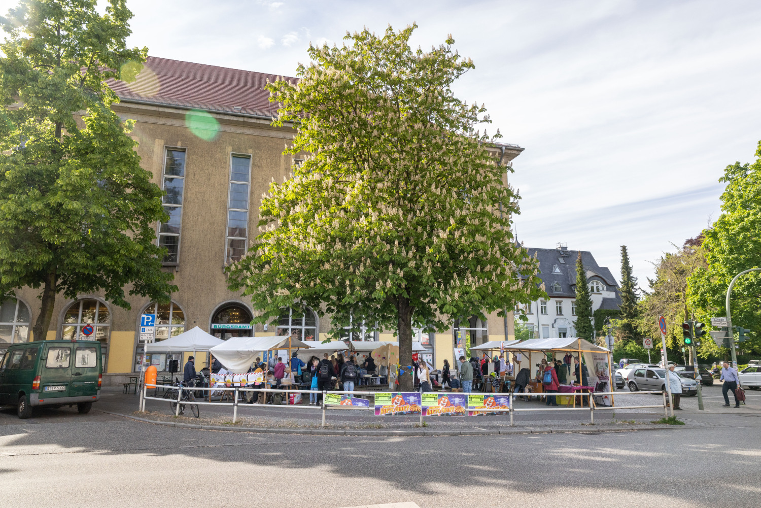 Blick von der anderen Straßenseite auf den Platz um die Kastanie vor dem Bürgeramt Steglitz-Zehlendorf, Teltower Damme / Ecke Kirchstraße. Die Sonne scheint, die Kastanie blüht.