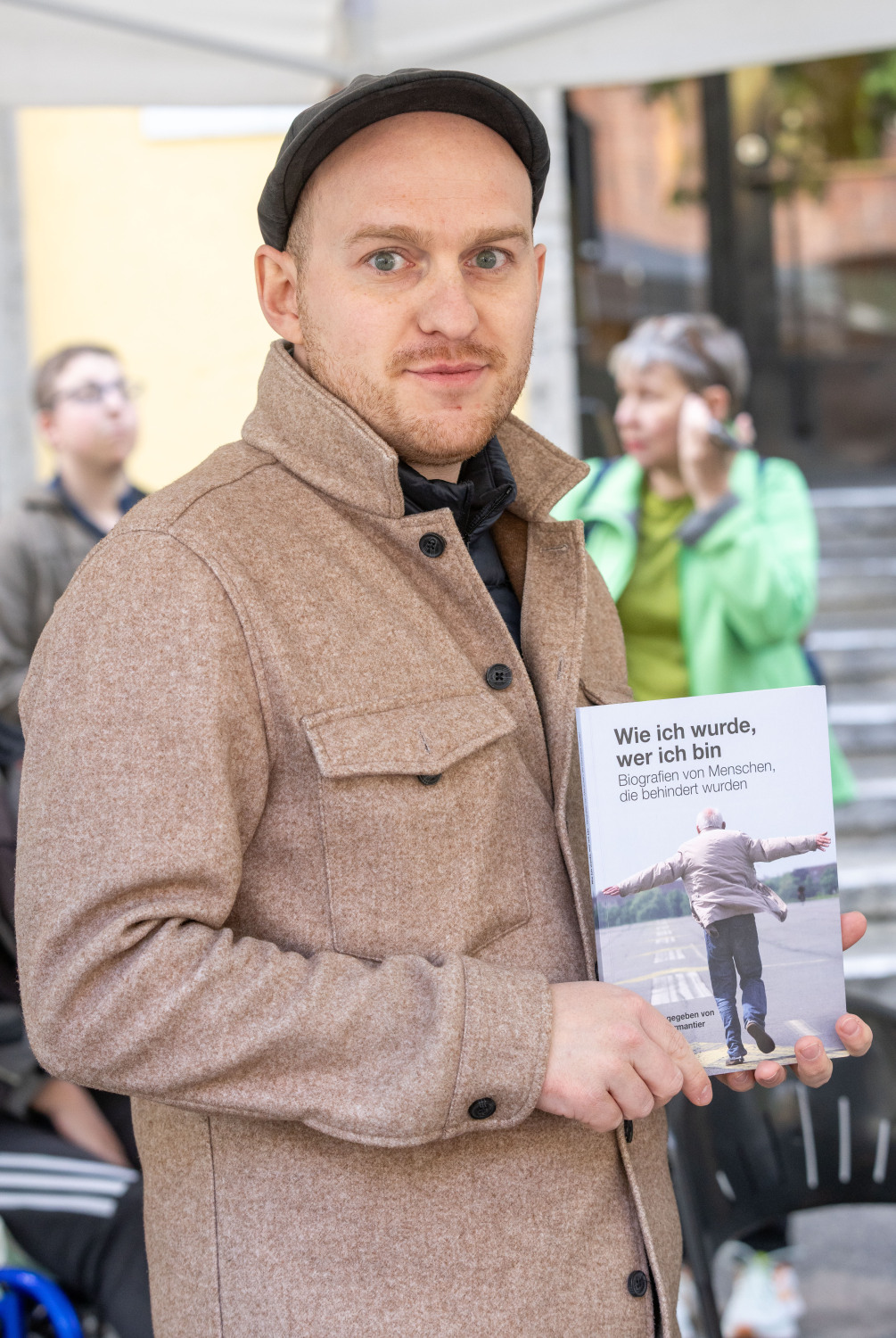 Eine Person in hellbrauner Jacke und mit brauner Mütze hält das Buch "Wie ich wurde wer ich bin." von der Zukunftssicherung Berlin e. V.