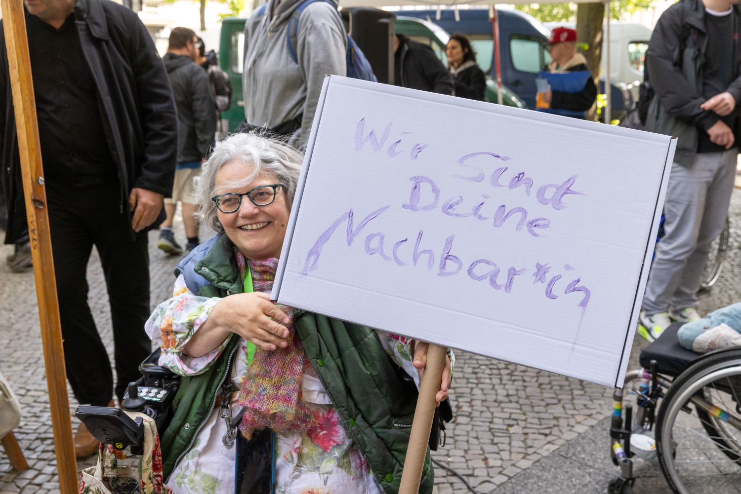 Die Beauftragte für Menschen mit Behinderungen von Steglitz-Zehlendorf, Eileen Moritz, mit ihrem Demoschild "Wir sind Deine Nachbar*in" in lilafarbener Schrift. Frau Moritz lacht
