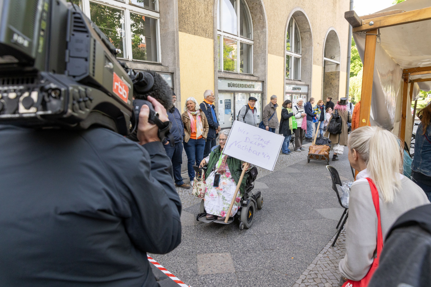 Der rbb nimmt mit der Fernsehkamera die Menschenrechtskette auf und Frau Moritz, die Beauftragte für Menschen mit Behinderungen, saust mit ihrem Rolli und einem Demoschild in der Hand durchs Bild. Auf dem Schild steht: Wir sind Deine Nachbar*in. Sie lacht in die Kamera.