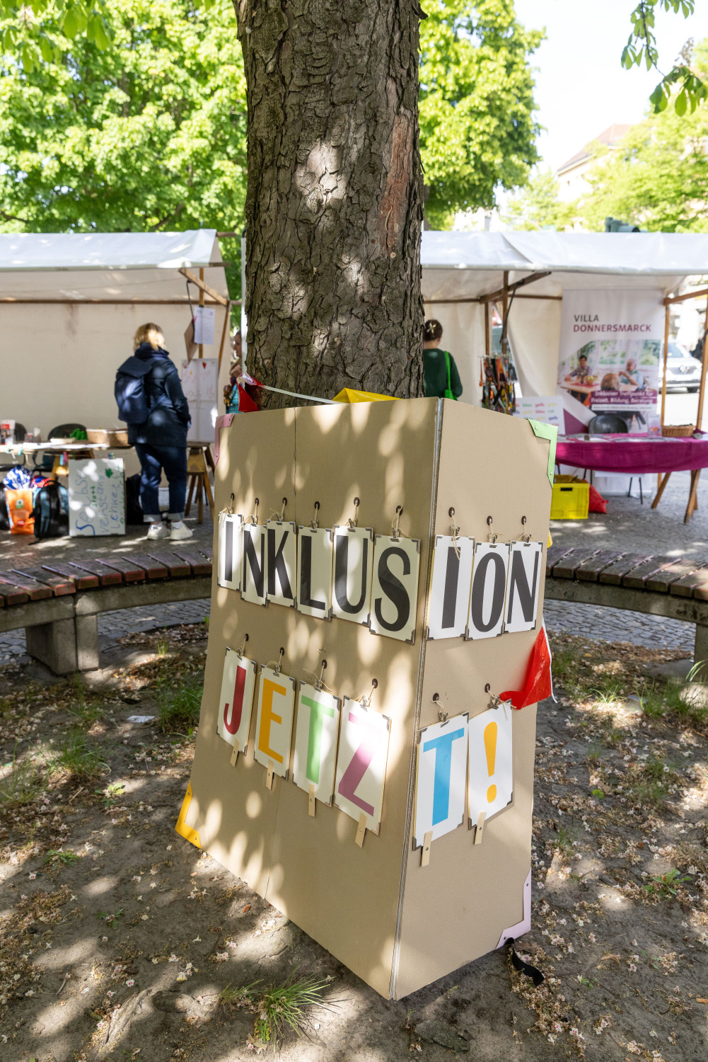 Ein riesiges Pappschild auf dem in Großbuchstaben INKLUSION JETZT! steht, wurde um die Kastanie gewickelt. Das Wort INKLUSION hat schwarze Buchstaben, das Wort JETZT! hat verschiedene bunte Buchstaben.