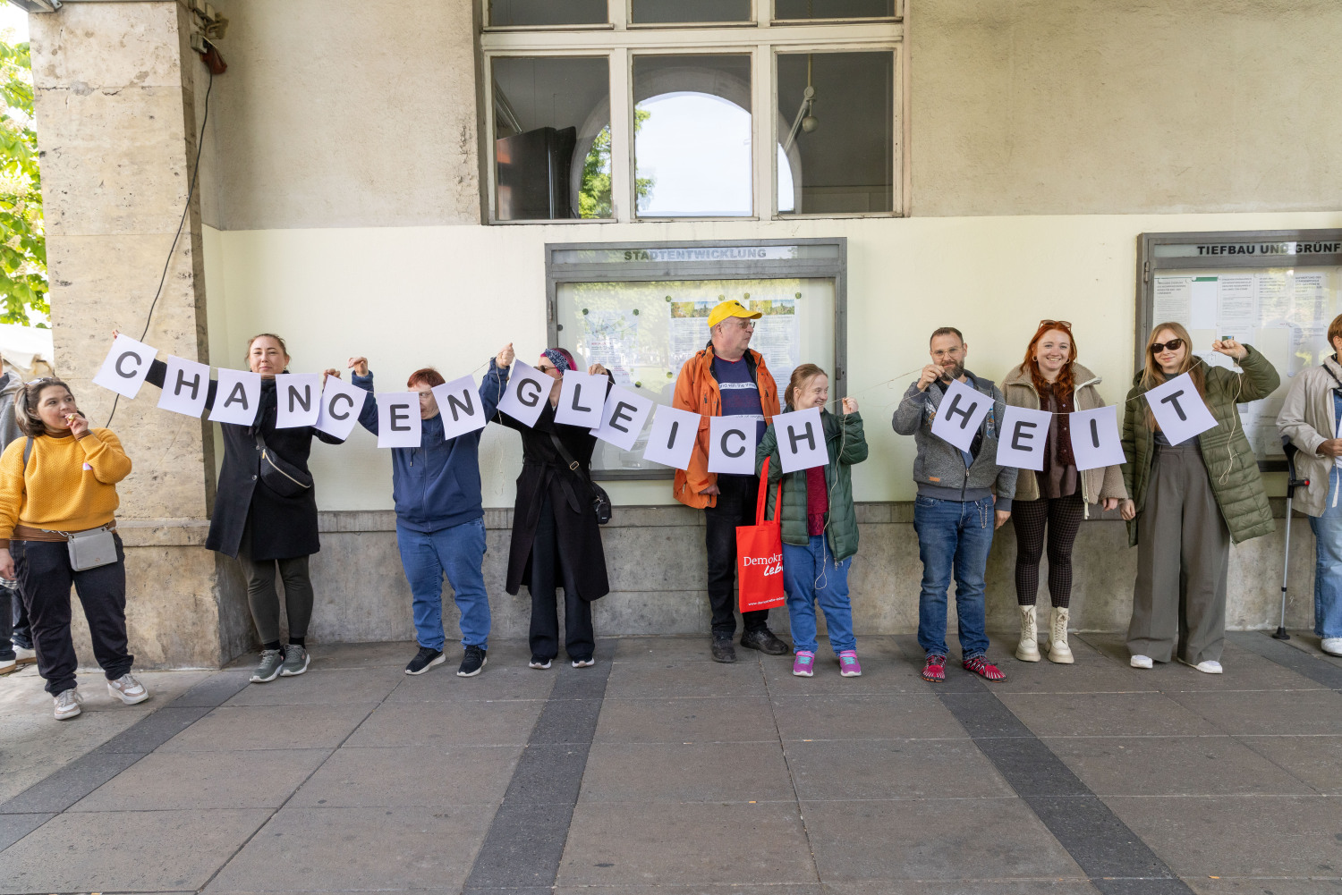 Ein Teil der Menschenrechtskette. Mehrere Personen halten eine lilafarbene Girlande fest. Diese ergibt das Wort CHANCENGLEICHHEIT. Die Menschen machen Lärm.