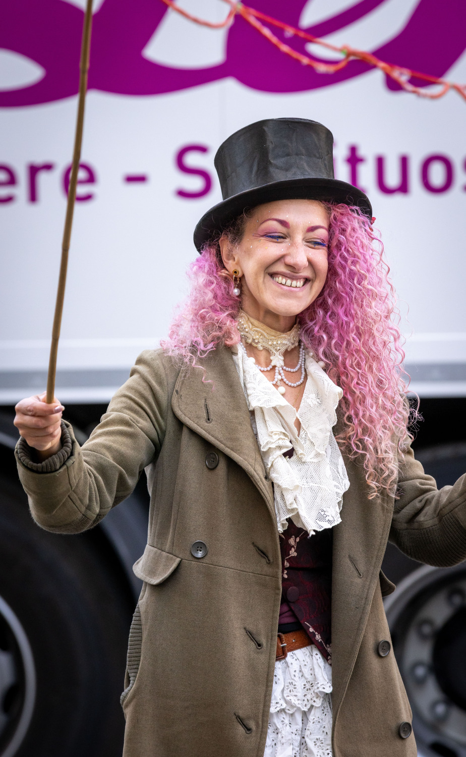 Corinne von Bubbles Powder. Sie trägt einen braunen Frack, eine weiße Bluse mit Rüschen, eine kleine schwarze Weste, einen schwarzen Zylinder und sie hat rosafarbene Haare. Sie lacht.