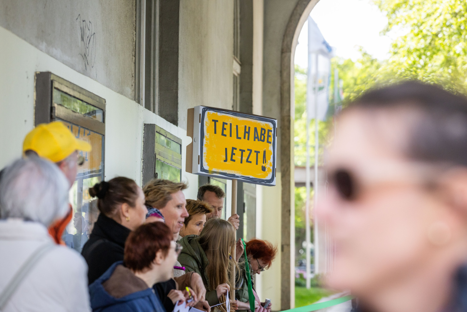 Ein Blick auf die Menschenkette unter den Arkaden des Rathauses. Jemand hält ein Schild. Es ist gelb, hat einen schwarzen Rand und schwarze Schrift TEILHABE JETZT!