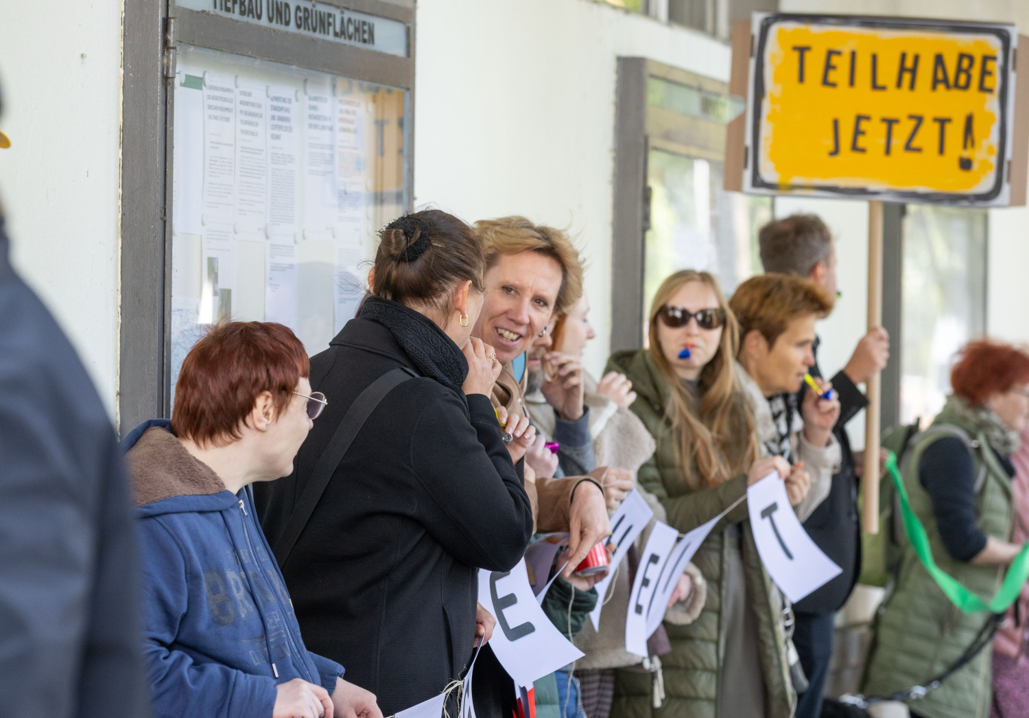 Ein Teil der Menschenrechtskette. Mehrere Personen halten einen Teil der lilafarbene Girlande fest, andere ein grünes Band, eine Person hält ein Demoschild auf dem steht TEILHABE JETZT! Es hat einen gelben Hintergrund, ist schwarz umrandet, die Schrift ist schwarz.