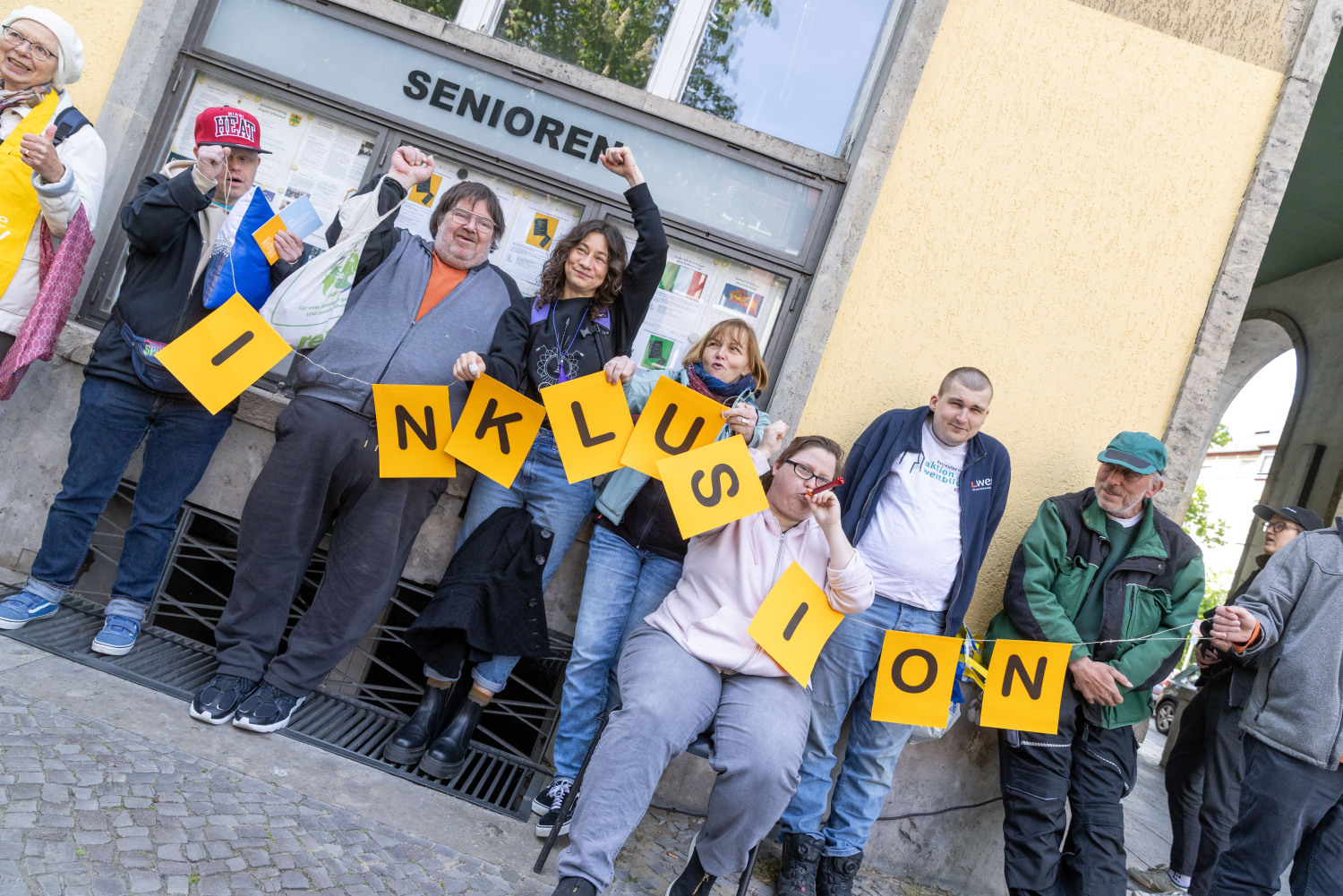 Die Menschenrechtskette. Verschiedene Personen halten eine orangefarbene Girlande. Darauf steht INKLUSION