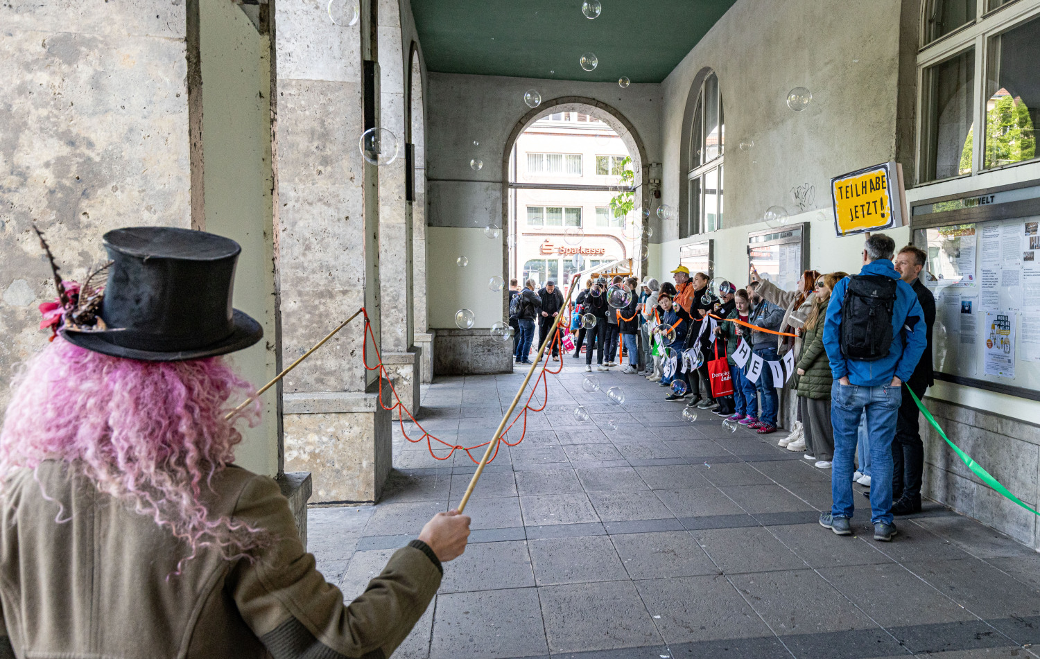 Ein Blick von weitem auf die Menschenkette unter den Arkaden des Rathauses. Die Menschen halten etwas in den Händen. Man erkennt orangefarbenes und ein grünes Band. Jemand hält ein Schild. Es ist gelb, hat einen schwarzen Rand und schwarze Schrift TEILHABE JETZT! Im Vordergrund sieht man eine Frau von hinten. Sie hat rosa Haare, einen schwarzen Zylinder, eine bräunliche Jacke und macht scheinbar riesige Seifenblasen.