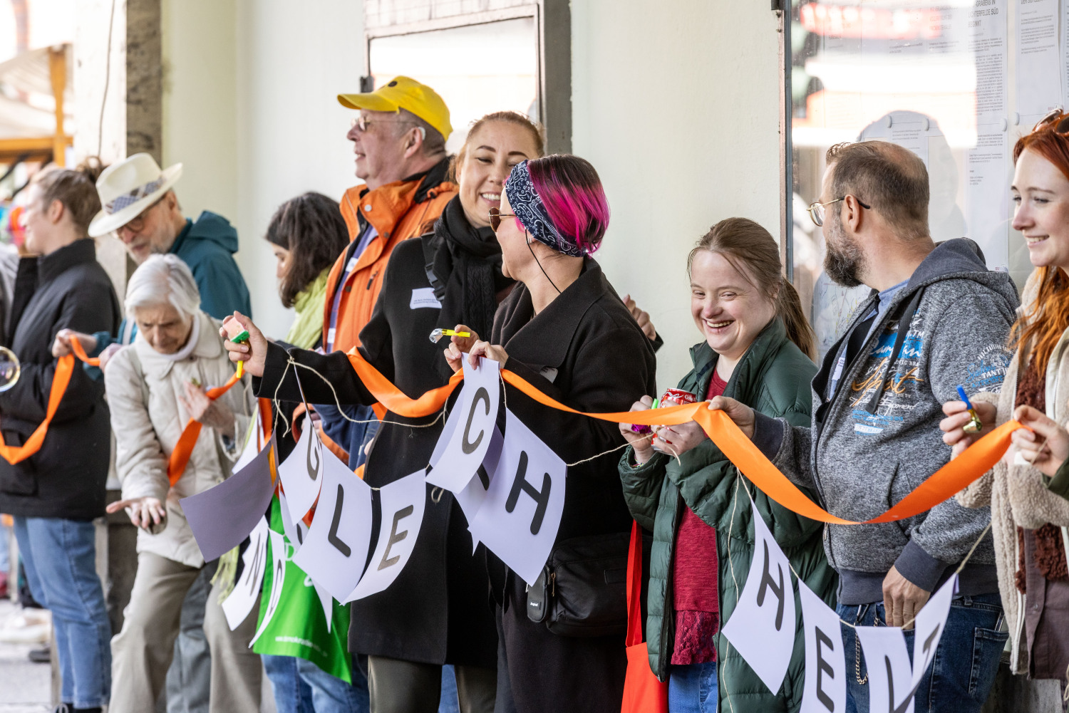 Ein Blick auf die Menschenkette. Die Menschen halten eine Girlande mit lilafarbenen Blättern an einem orangefarbenen Band. Die Menschen sind alle verschieden. Ein Teil der Girlande lautet: Gleichheit