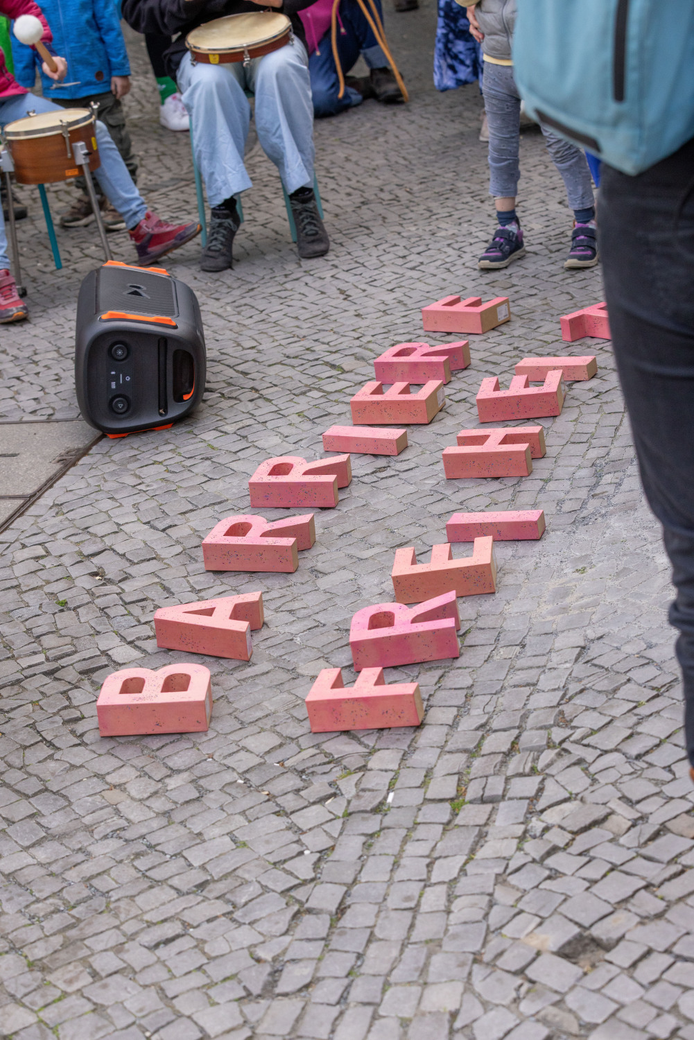Auf Kopfsteinpflaster liegen neben einer Lautsprecherbox große rosafarbene Buchstaben. Sie ergeben das Wort BARRIEREFREIHEIT.