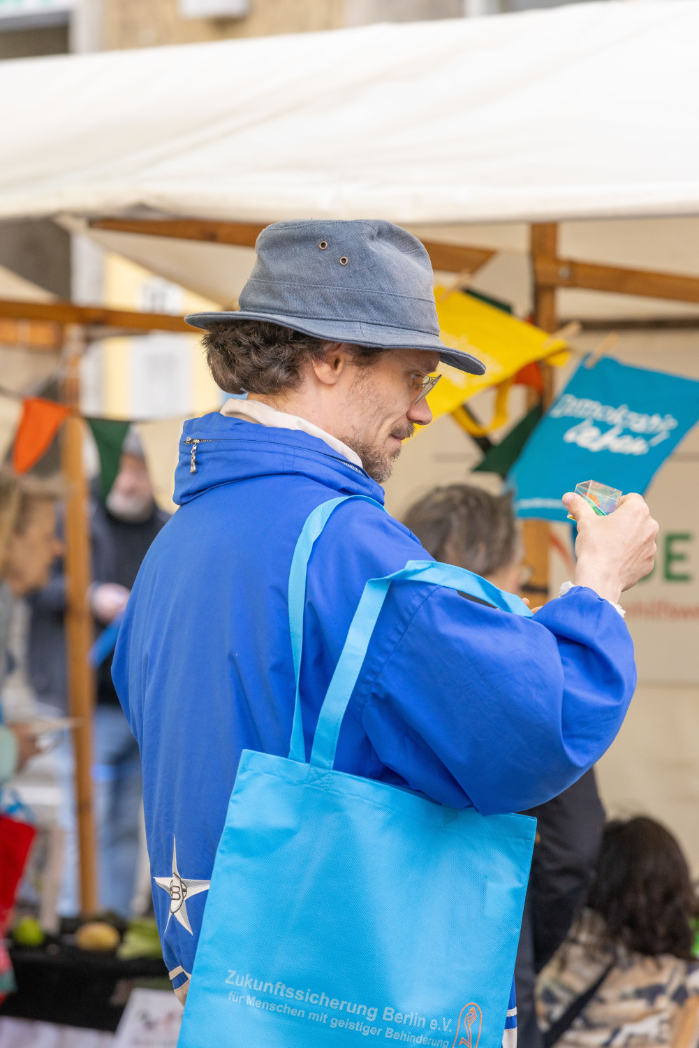 Eine Person mit einer blauen Jacke, einem blaugrauen Hut und einem leuchtend blauen Beutel der Zukunftssicherung steht an einem Stand. Im Hintergrund ist ein blauer Wimpel mit weißer Schrift Demokratie leben. Eine schöne Farbzusammenstellung.