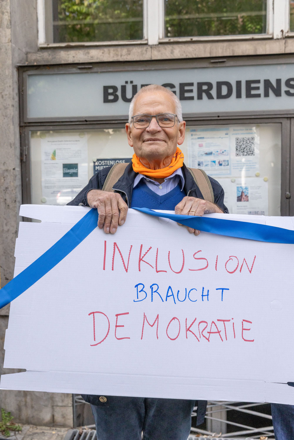 Ein Mitglied des Beirates für Menschen mit Behinderungen hält ein blaues Band zusammen mit einem Demoschild auf dem in roter und blauer Schrift stehtt "INKLUSION braucht DEMOKRATIE". Er trägt ein blaues Hemd und eine orangefarbenes Halstuch.