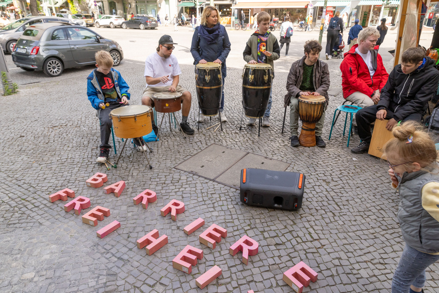 Auf dem Bild sind Mitglieder der Trommelgruppe vom DRK. Sie sitzen im Halbkreis und trommeln. Der Halbkreis wird durch große rosafarbene Buchstaben geschlossen. Sie ergeben das Wort BARRIEREFREIHEIT.