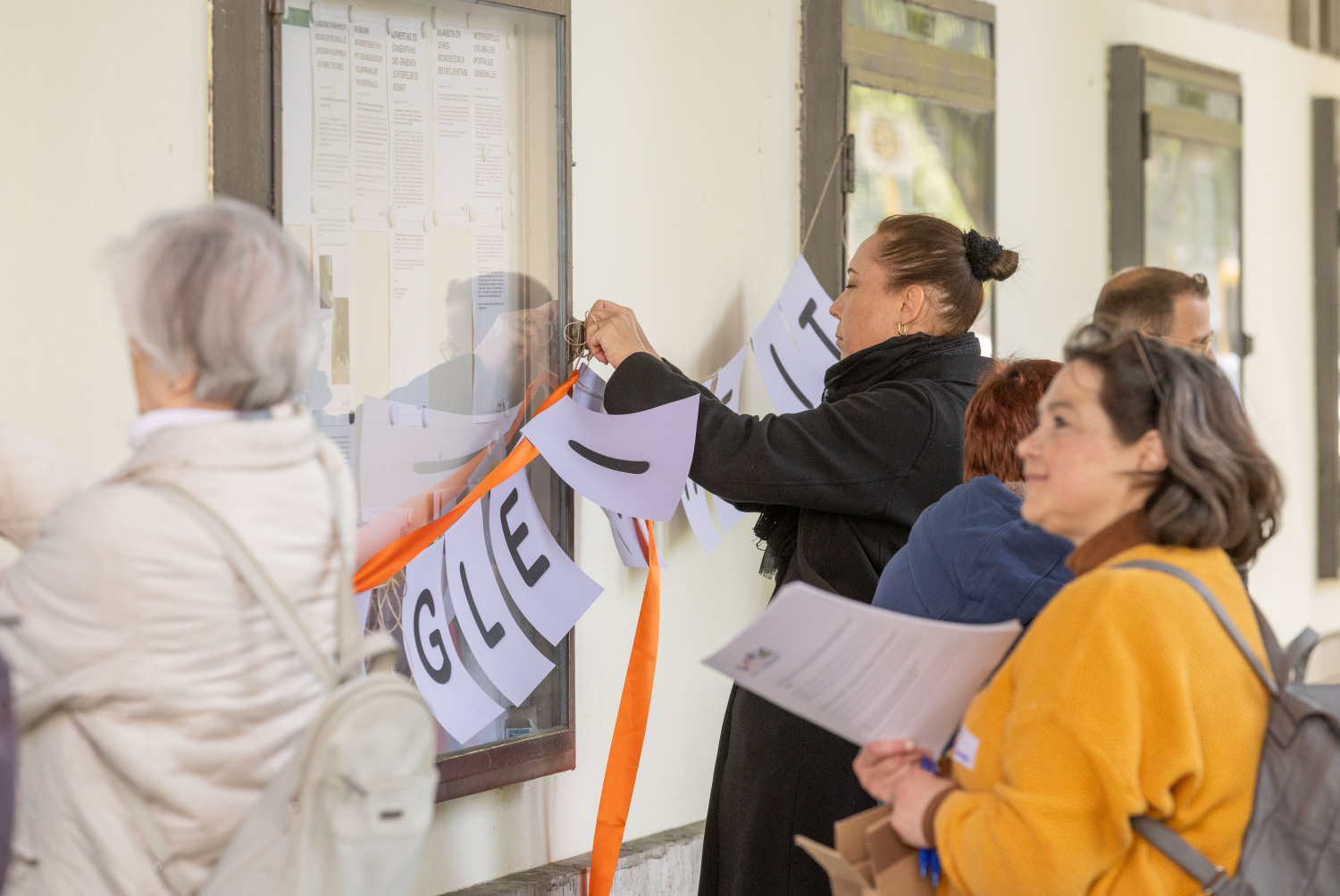 Menschen versuchen die lilafarbene Girlande Chancengleichheit und ein orangefarbenes Band an Glasschaukästen zu befestigen. Eine Person schaut lächelnd zu. Sie trägt einen leuchtenden curryfarbenen Pullover.