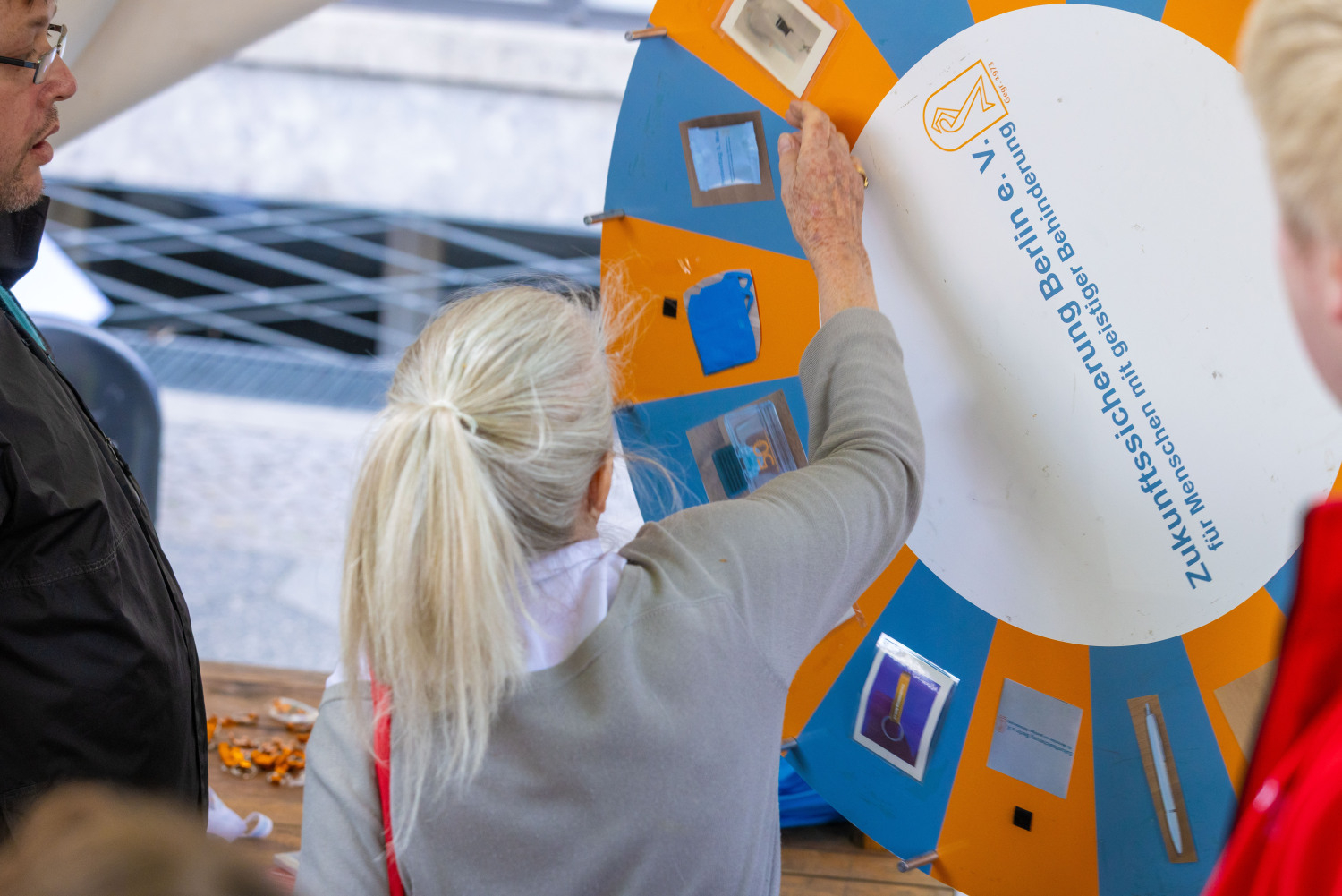 Eine Person mit grauem Zopf und grauer Kleidung dreht an einem Glücksrad. Das Glücksrad hat blaue und orangefarbene Felder in der Mitte einen großen weißen Kreis mit blauer Aufschrift Zukunftssicherung Berlin e. V. für Menschen mit geistiger Behinderung
