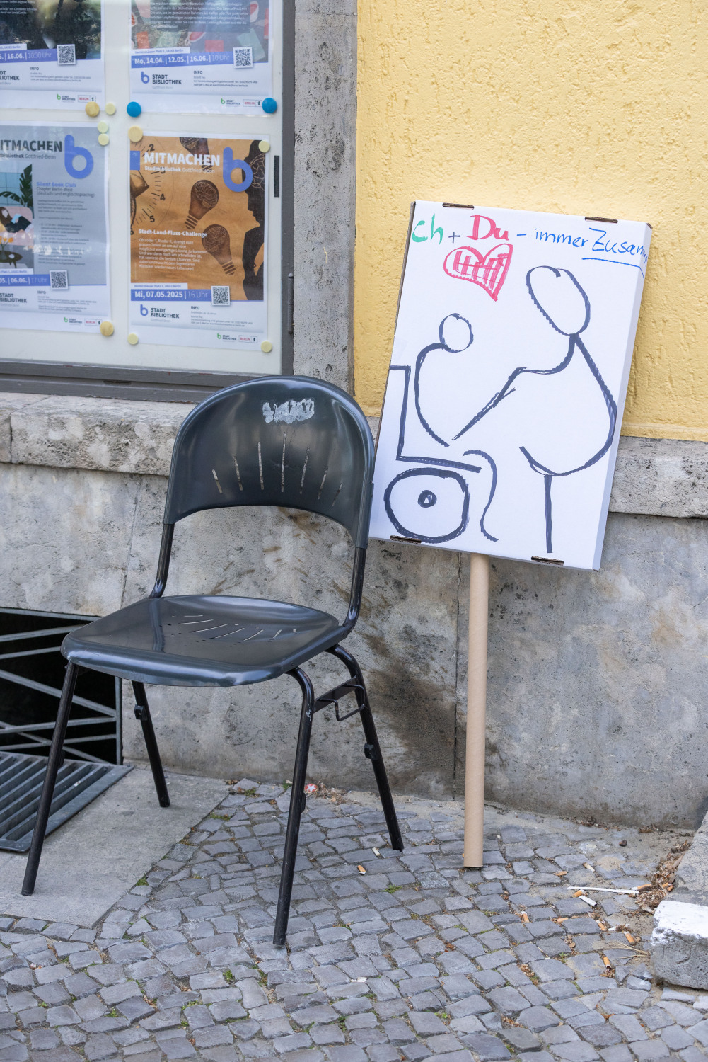 Ein Demoschild lehnt neben einem schwarzen Stuhl an einer gelblichen Hauswand. Auf dem Schild steht in grüner, roter und blauer Schrift "Ich + Du = Immer Zusammen". Es wurde ein abstraktes Bild/Symbol dazu gemalt: Kind im Rolli und Mutter halten sich an den Händen.
