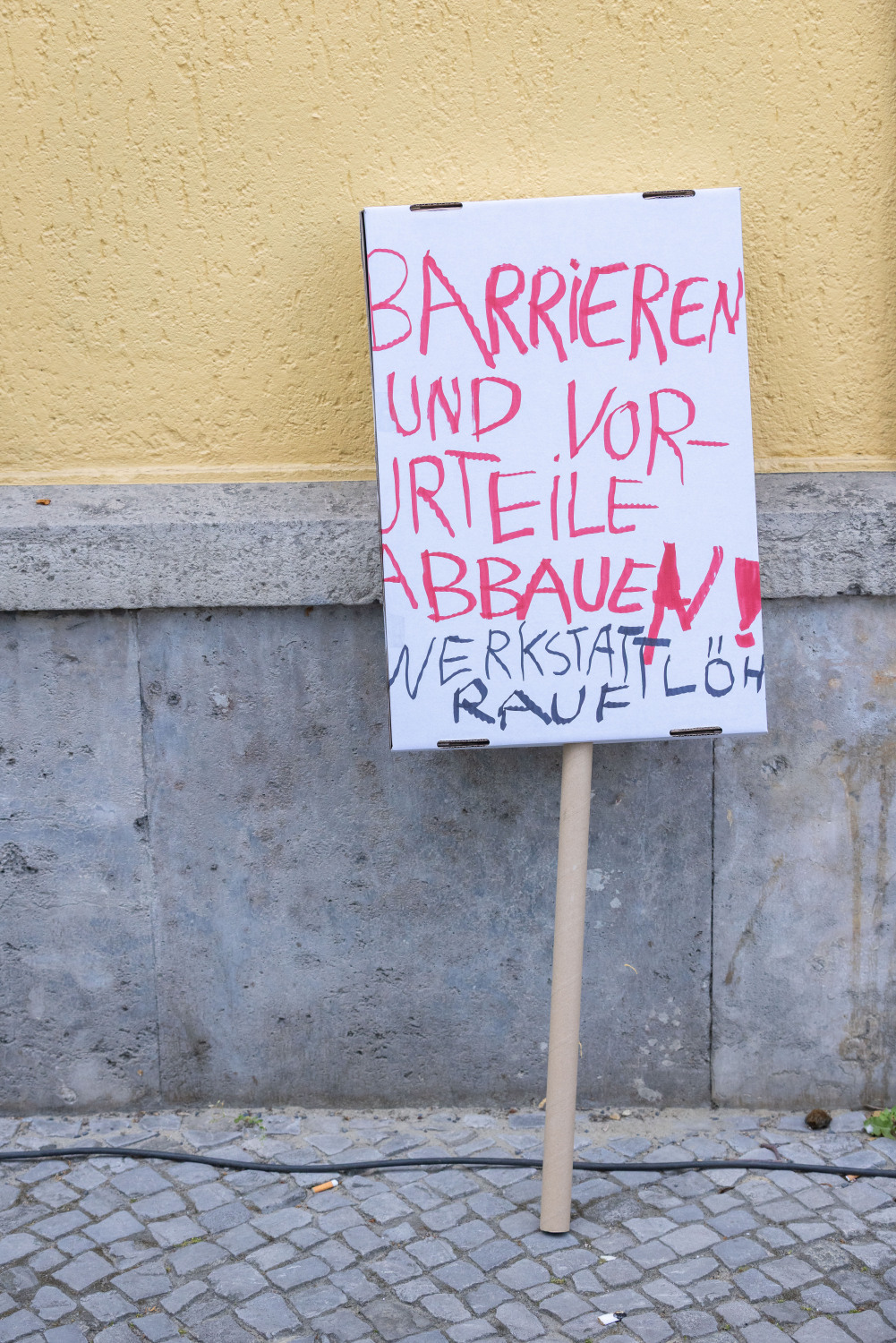 Ein Demoschild lehnt an einer Hauswand. Die Wand ist oben gelb und unten grau. Auf dem Schild steht in roter Schrift "Barrieren und Vorurteile abbauen" und in schwarzer Schrift "Werkstattlöhne rauf".