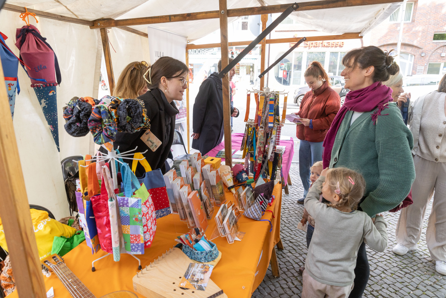 Eine Person mit Kind informiert sich an einem der Stände. Der Stand hat eine orangefarbene Tischdecke und alles ist sehr bunt. Im Hintergrund ist eine andere Person an einem anderen Stand zu sehen.