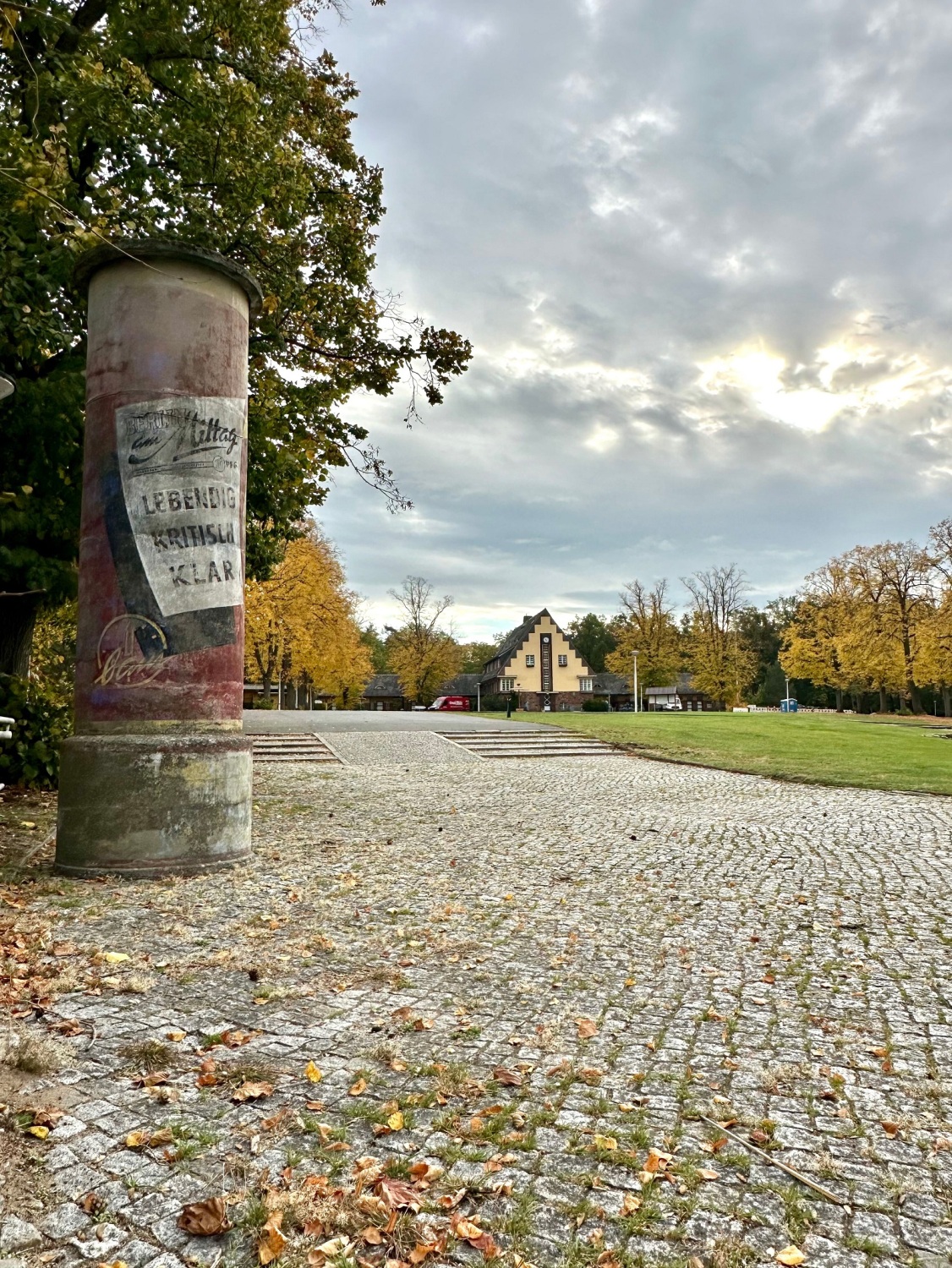 Litfaßsäule mit historischem Werbeplakat vorne und das Hauptgebäude des Strandbads im Hintergrund