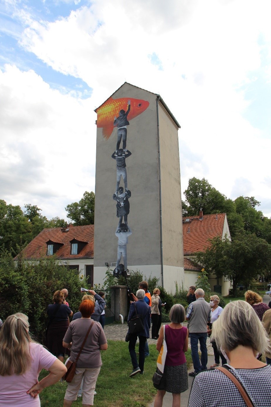 Eine Menschengruppe steht mit Abstand zueinander vor einem Turm. Auf dem Gebäude ist ein Bild von einer vier Mann starken Räuberleiter zu sehen. Die Person die ganz oben auf der Räuberleiter steht malt einen Goldfisch an die Wand.