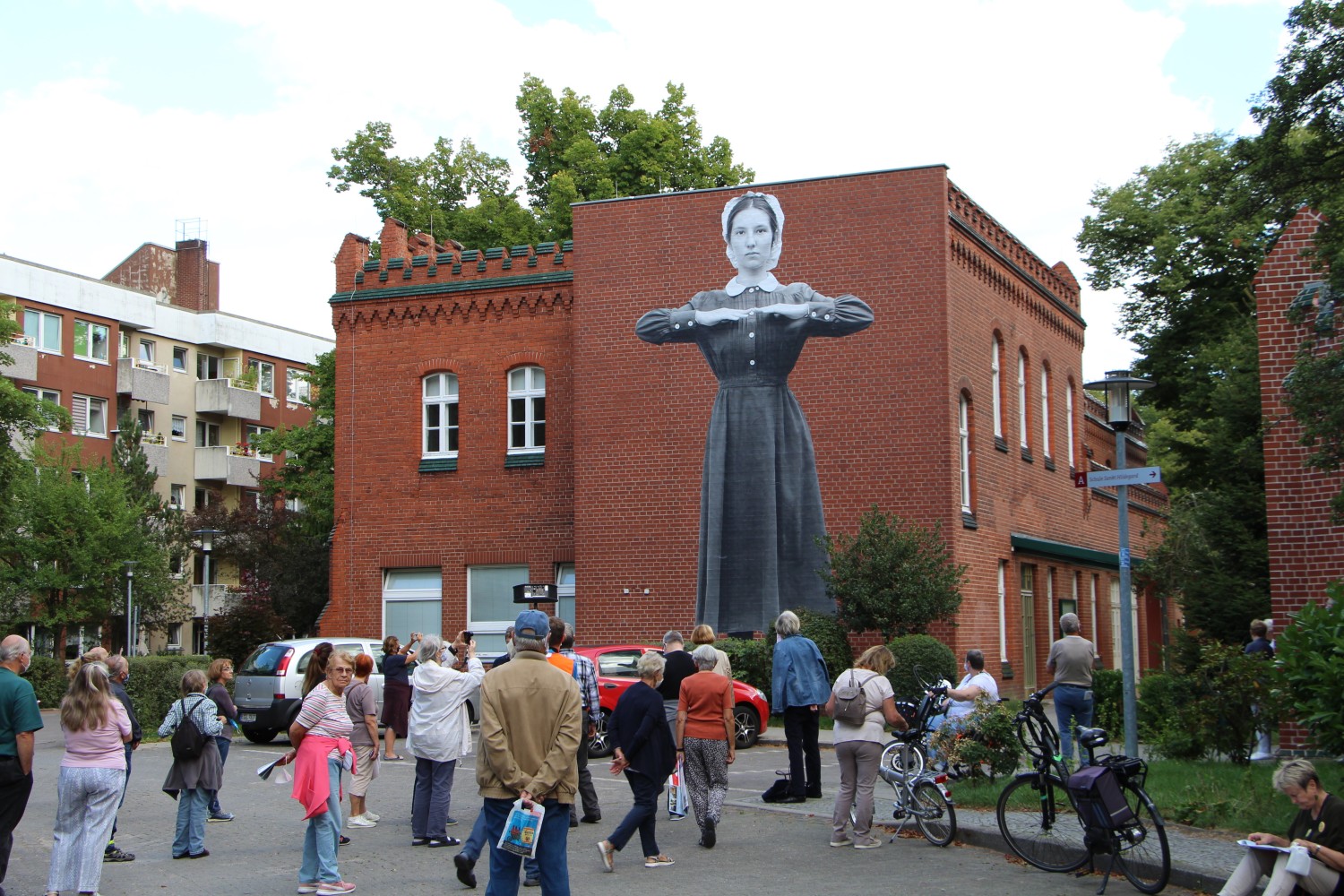 Eine Menschenmenge steht vor einem Gebäude. Auf der Hauswand ist eine Klosterfrau mit vor der Brust haltenden Unterarmen zu sehen.