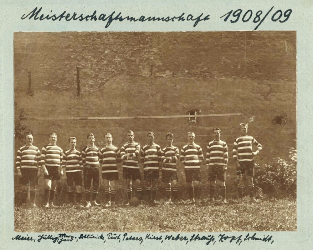 Schwarzweißfoto der nebeneinander vor einer Hauswand stehenden 11 Fußballer in gestreiften Trikots, handschriftlich sind die Namen der Spieler am unteren Bildrand zu hinzugefügt. 