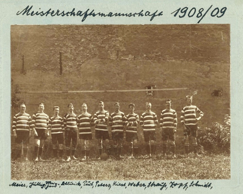 Schwarzweißfoto der nebeneinander vor einer Hauswand stehenden 11 Fußballer in gestreiften Trikots, handschriftlich sind die Namen der Spieler am unteren Bildrand zu hinzugefügt.