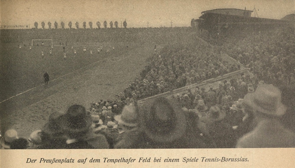 Ein Blick über die Tribüne zeigt viele Zuschauer mit Hüten von hinten, links im Hintergrund ist das Spielfeld zu sehen, auf dem das Spiel läuft. Bildunterschrift aus der Mannschaftszeitschrift: "Der Preußenplatz auf dem Tempelhofer Feld bei einem Spiele Tennis-Borussias."