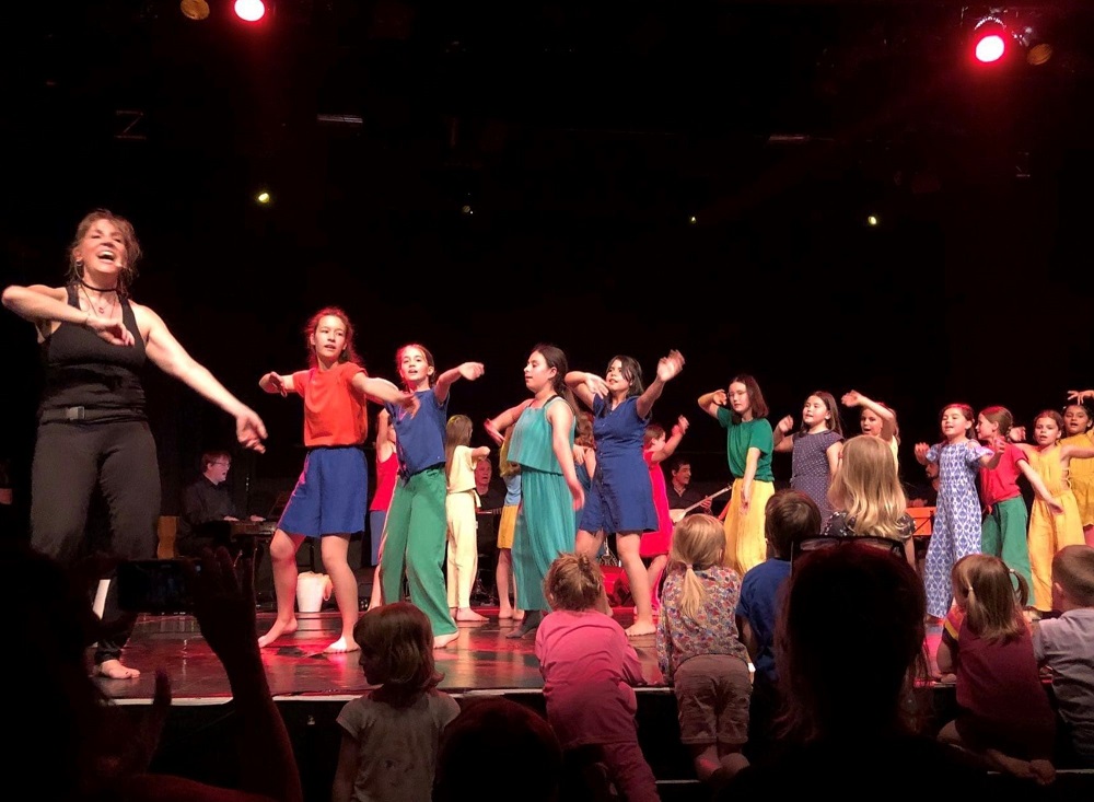 Eine Frau und Kinder singen und tanzen auf einer Bühne.