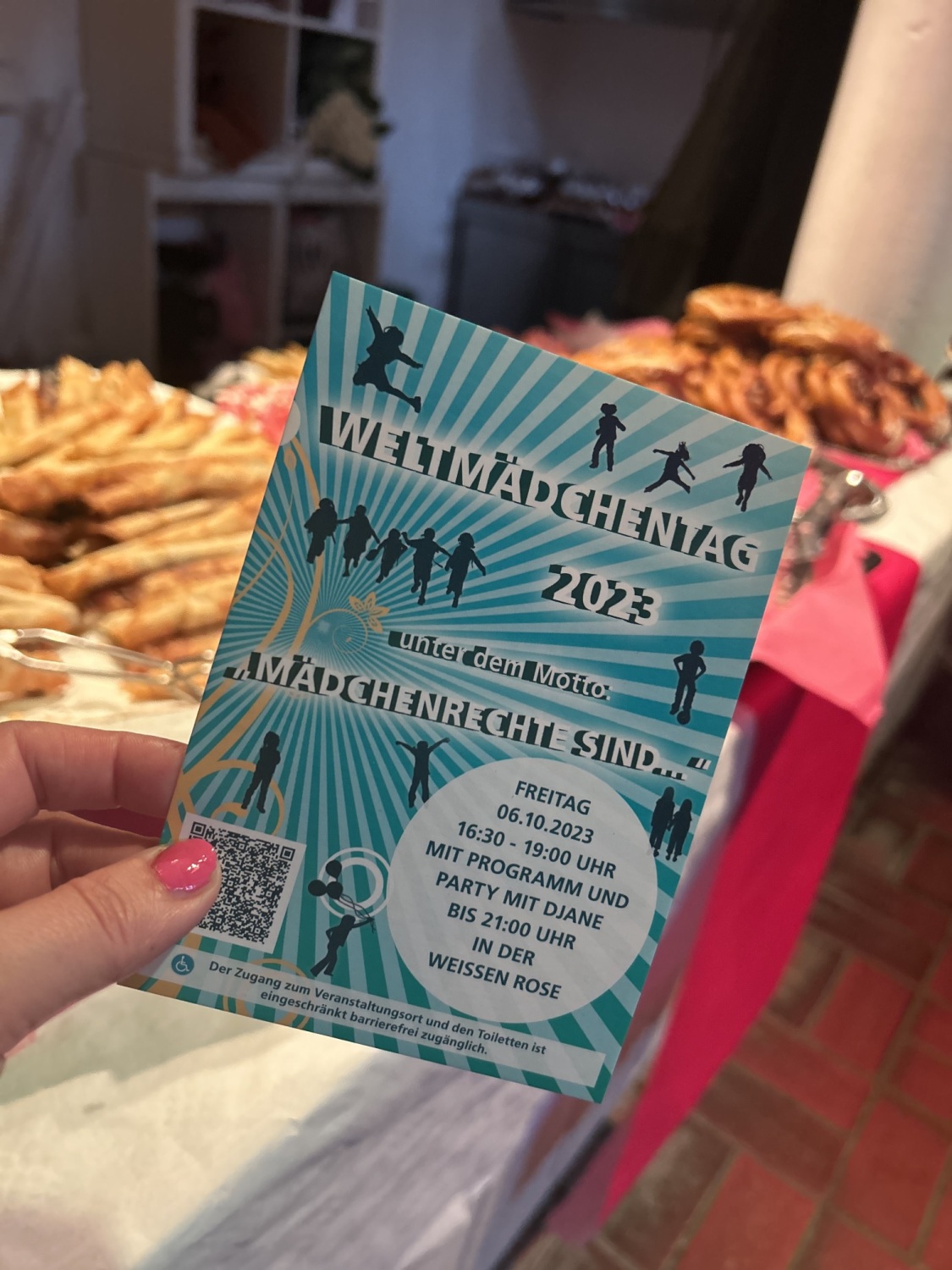 Ein Flyer zum Weltmächentag 2023 in der Weißen Rose wird hoch gehalten. Im Hintergrund ist ein Buffet mit Bretzeln und anderem Gebäck.