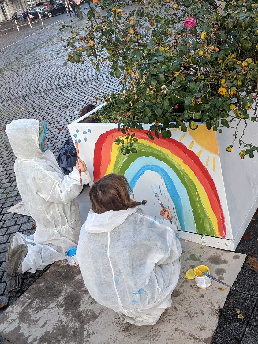 Kinder bemalen einen Blumenkübel mit einem Regenbogen