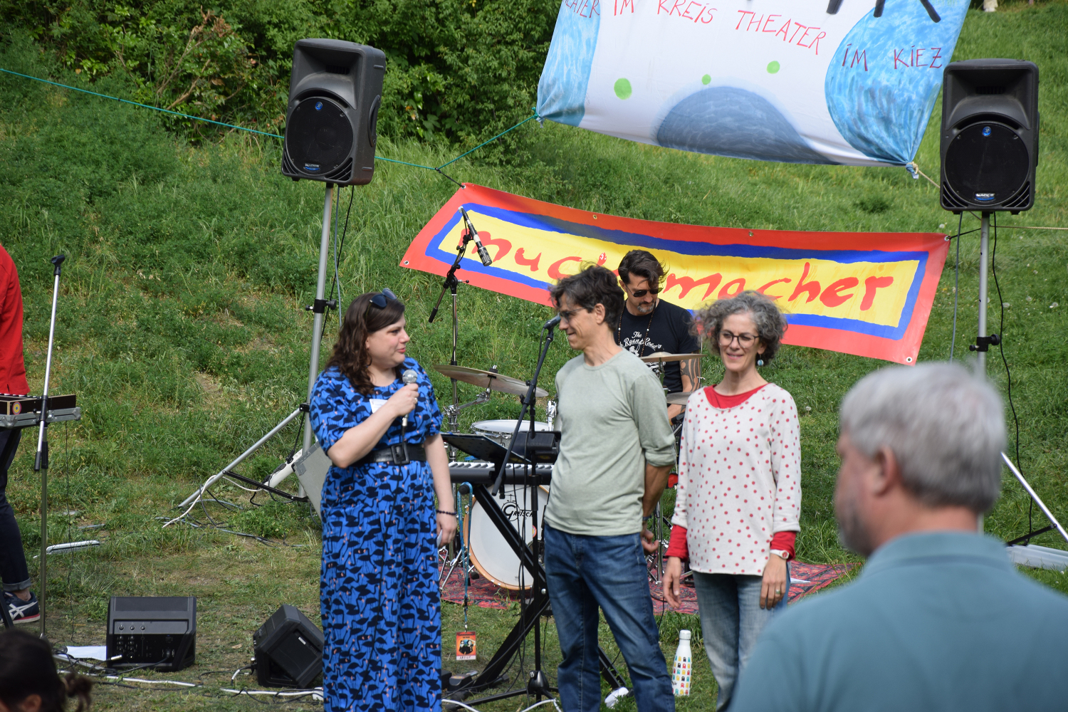 Drei Personen stehen auf einer Grünfläche an Mikrofonständern. Im Hintergrund sitzt jemand an einem Schlagzeug.
