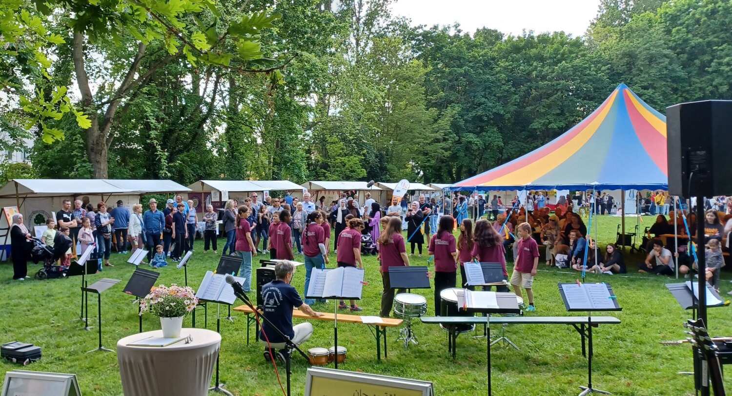 Auf einem Fest auf einer Grünfläche sind viele Menschen. In der Mitte stehen Kinder in einem Kreis.