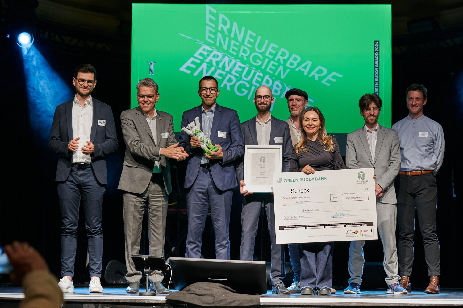 Auf einer Bühne stehen Personen zusammen und halten eine Bären-Statue, eine Urkunde und einen Scheck in den Händen.