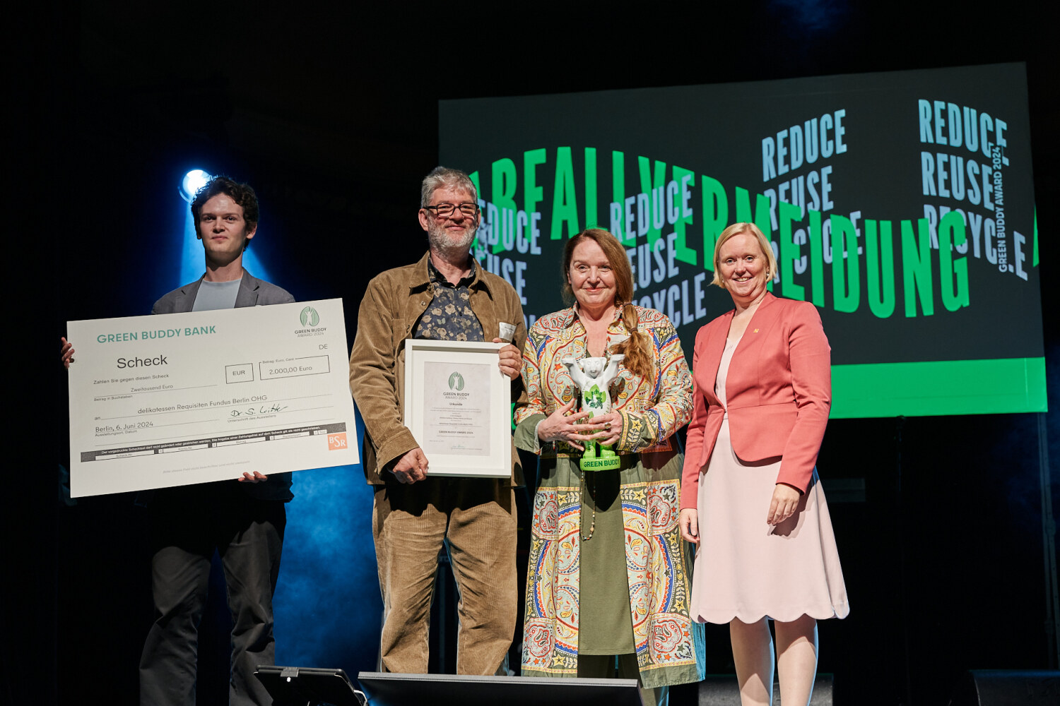 Auf einer Bühne stehen Personen zusammen und halten eine Bären-Statue, eine Urkunde und einen Scheck in den Händen.