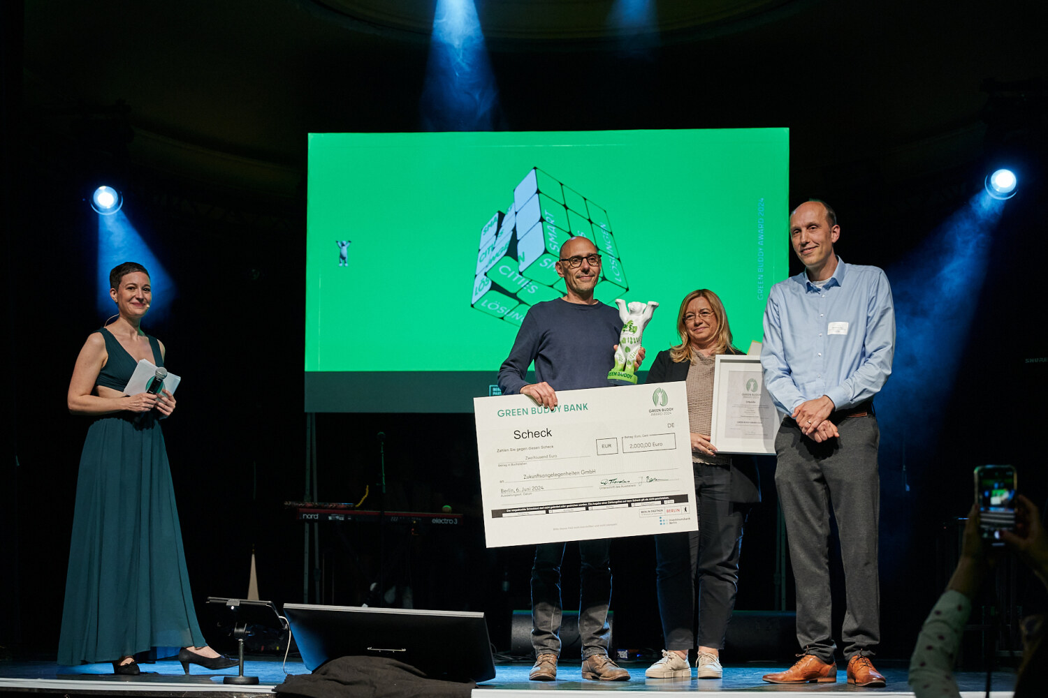 Auf einer Bühne stehen Personen zusammen und halten eine Bären-Statue, eine Urkunde und einen Scheck in den Händen.