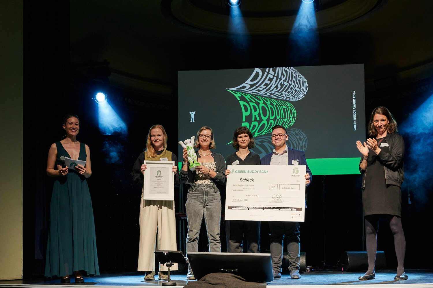 Auf einer Bühne stehen Personen zusammen und halten eine Bären-Statue, eine Urkunde und einen Scheck in den Händen.