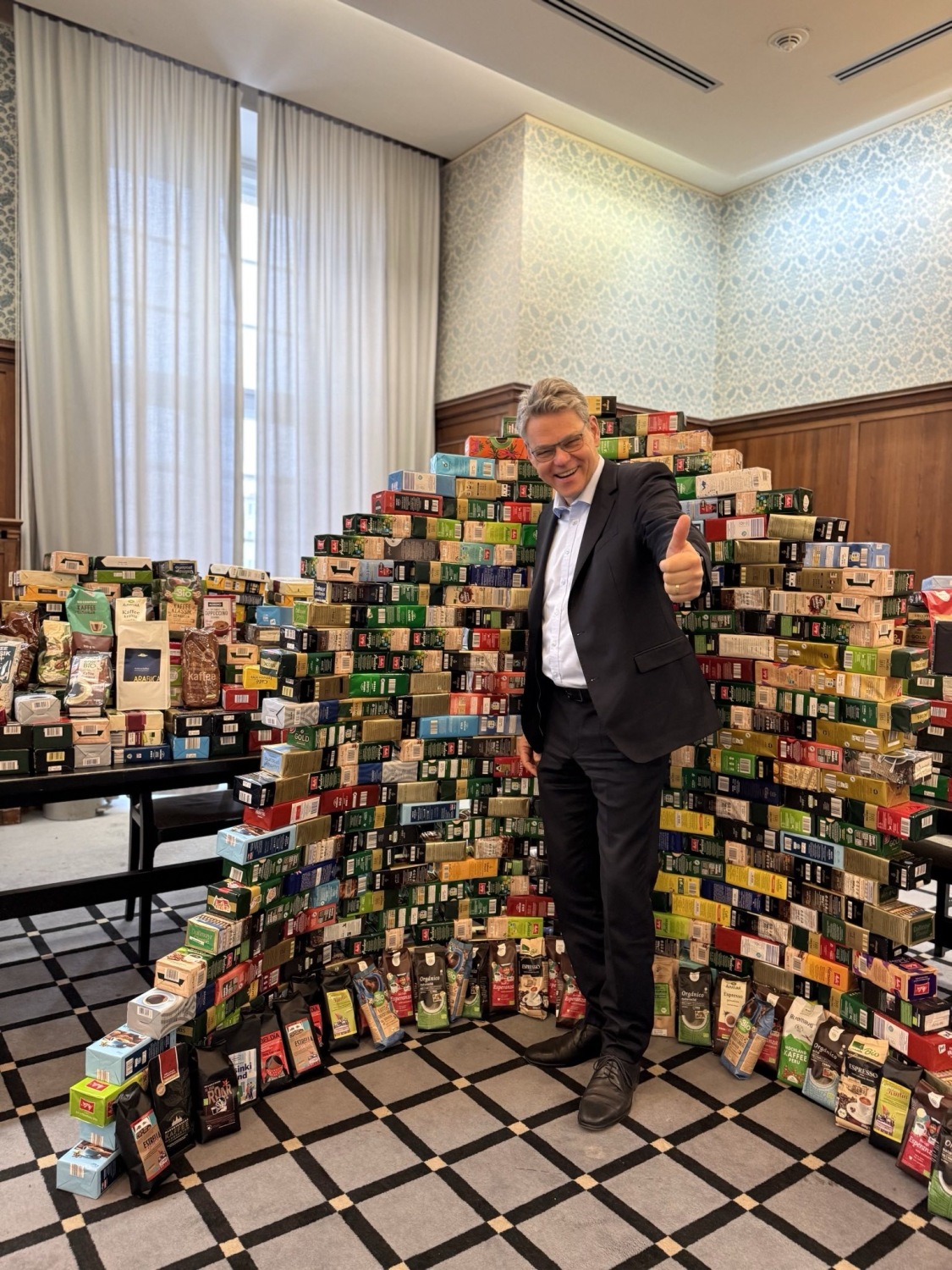 Ein Mann steht vor vielen Kaffeepäckchen und zeigt den Daumen hoch.