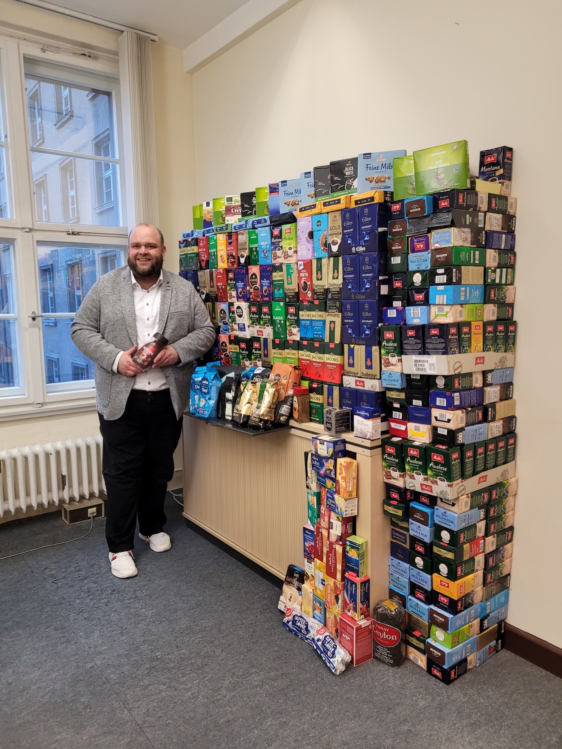 Ein Mann steht vor vielen Kaffeepäckchen und hält auch eines in der Hand.
