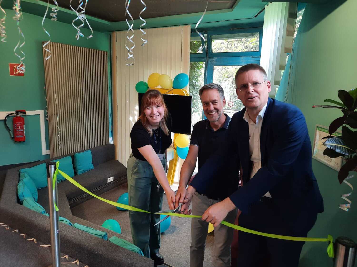 Drei Personen stehen hinter einem Band und schneiden es mit einer Schere durch. Sie stehen in einem geschmückten Raum mit Sitzmöglichkeiten und einem großen Bildschirm.