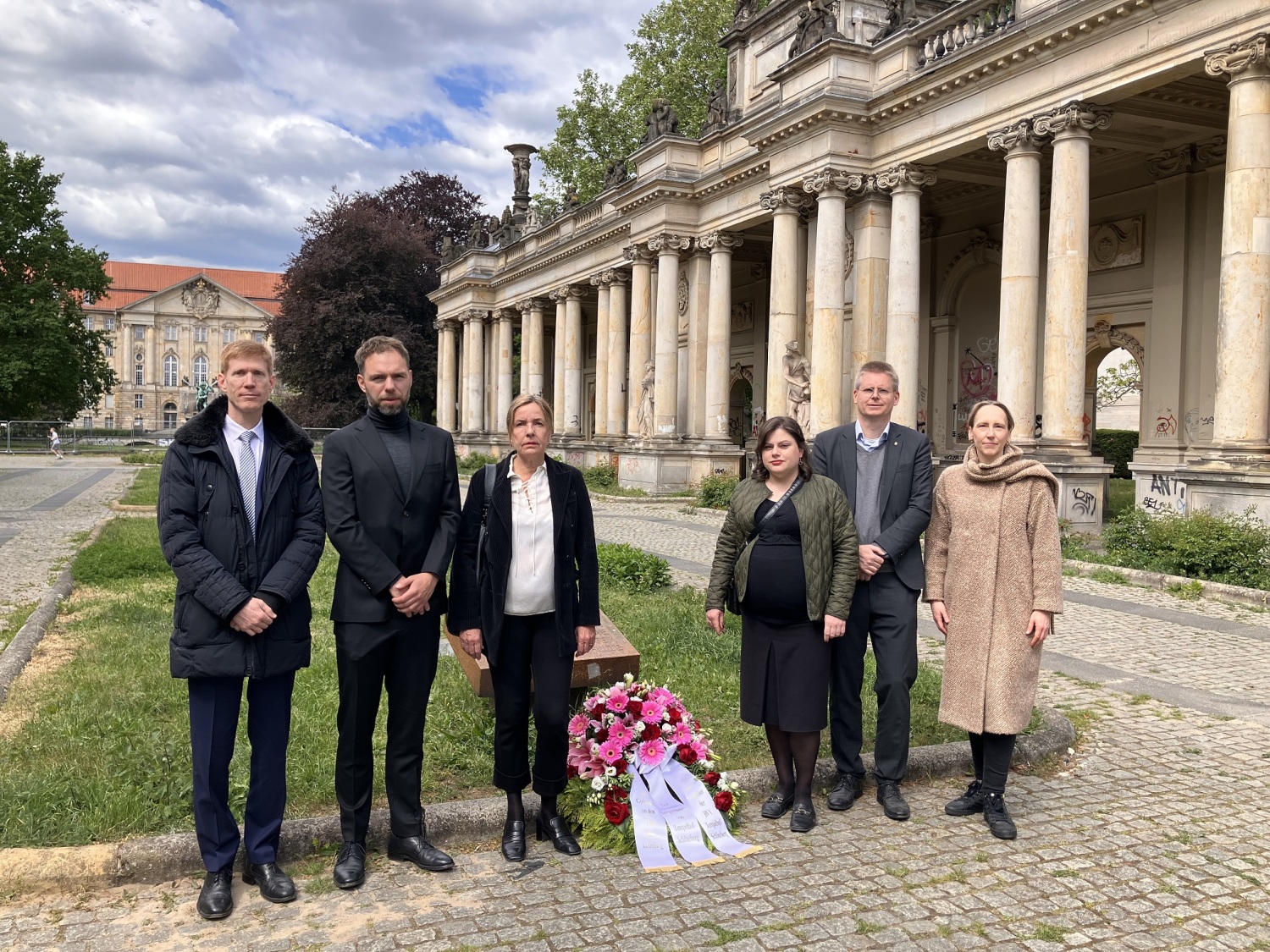 Sechs Personen stehen neben einem Kranz, der vor einem Gedenkstein niedergelegt ist.