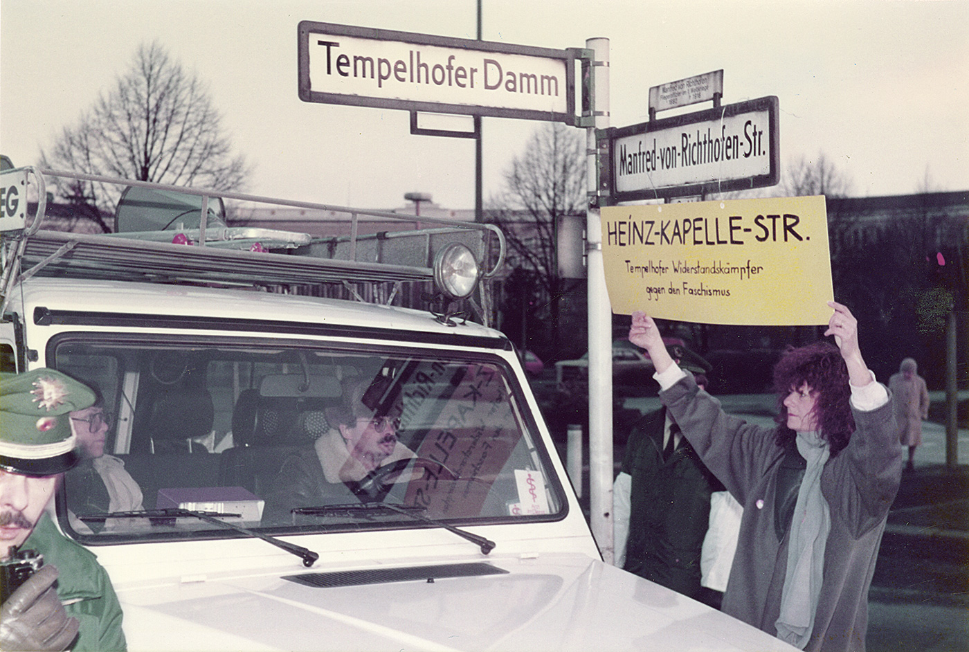 Eine Person hält ein Schild auf dem Hein-Kapelle-Straße steht unter das Straßenschild der Manfred-von-Richthofen-Straße.