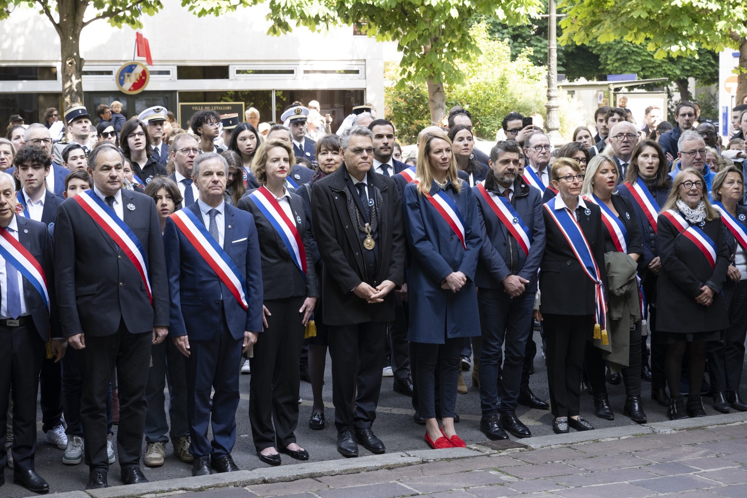 Viele Menschen stehen gaerade in Reihen auf einem Platz. Die meisten tragen eine Scherpe in den Farben der französischen Flagge.