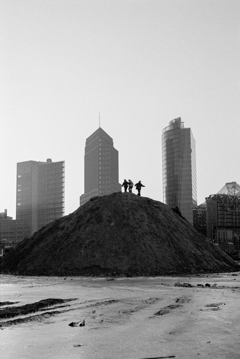 Schwarzweißfotografie von drei Menschen, die auf einem riesigen Erdhaufen stehen. Im Hintergrund sind drei Hochhäuser.