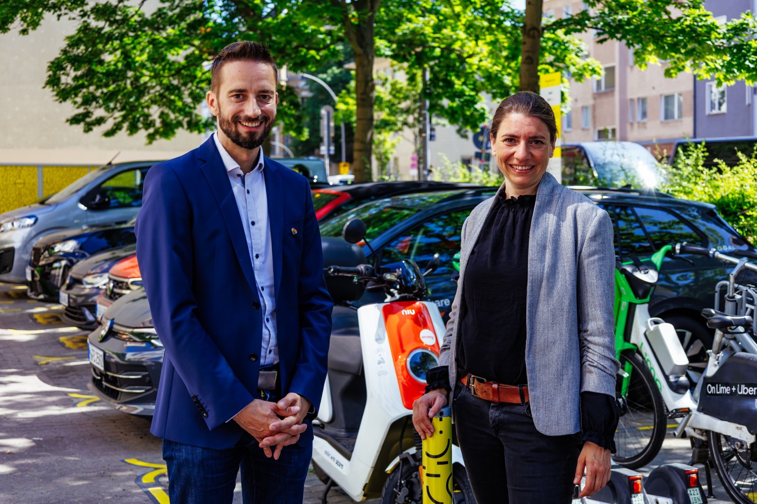 Zwei Personen stehen nebeneinander auf einem Platz mit Autos, Scootern, Rollern und Fahrrädern.